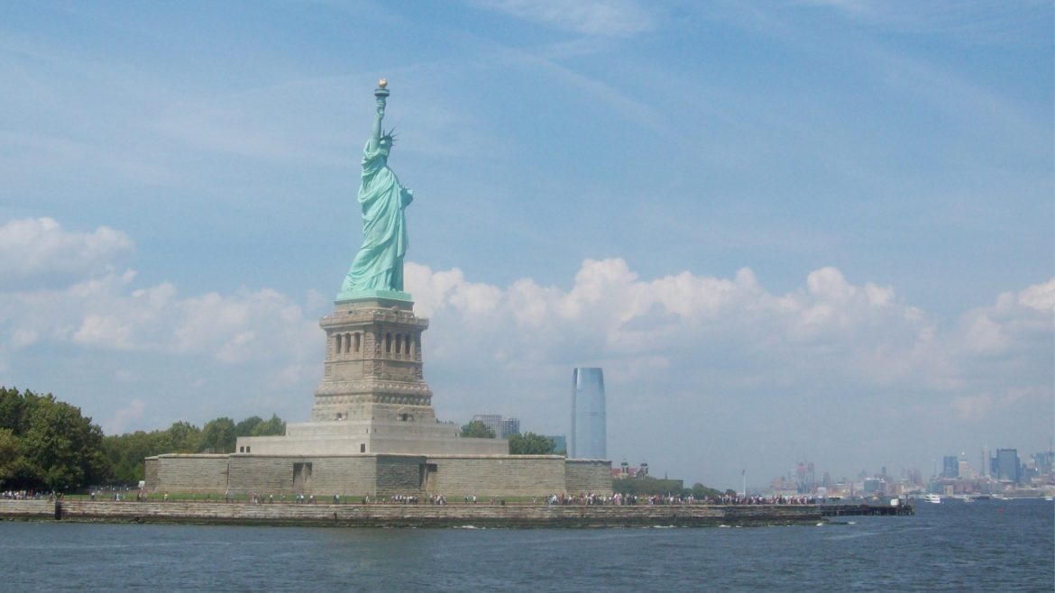 Side view of the Statue of Liberty standing on Liberty Island with the New York City skyline in the distance.